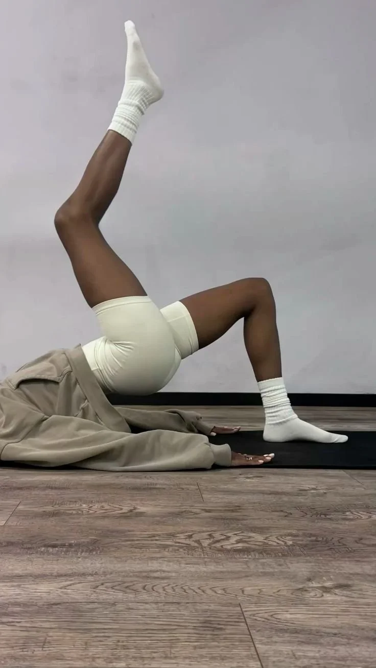 A person performing a yoga pose on a black mat against a plain white wall, with one leg extended upward and the other bent on the floor, wearing beige shorts, white socks, and a beige jacket.