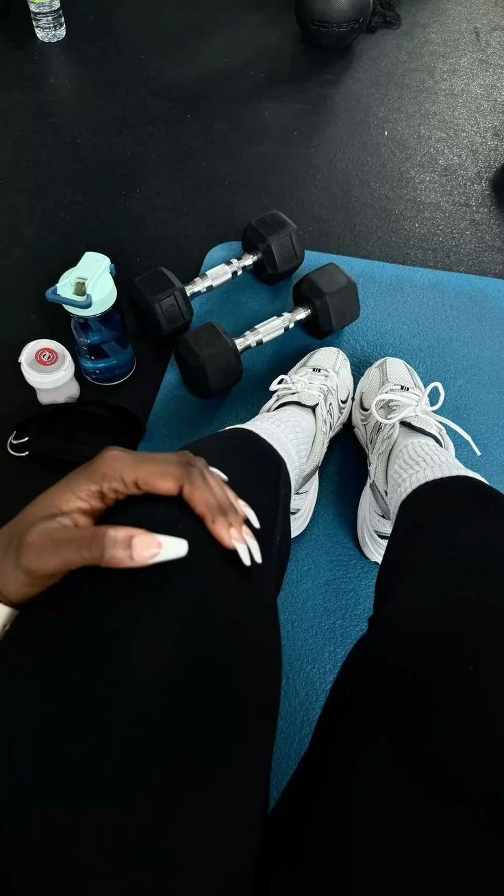 A person sitting on a gym mat with white athletic shoes, white socks, black leggings, and their hand resting on their knee. Nearby, there are two dumbbells, a blue water bottle, a small white container, and a light blue shaker bottle. The gym floor is black.