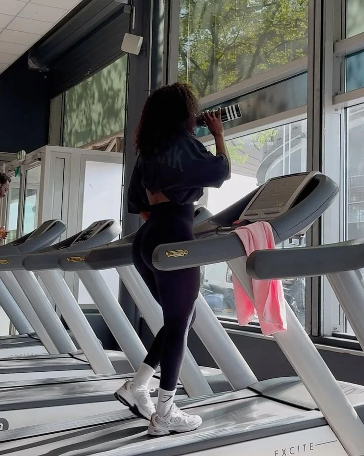 A woman with curly hair running on a treadmill next to large windows at a gym, with a pink towel draped over the treadmill