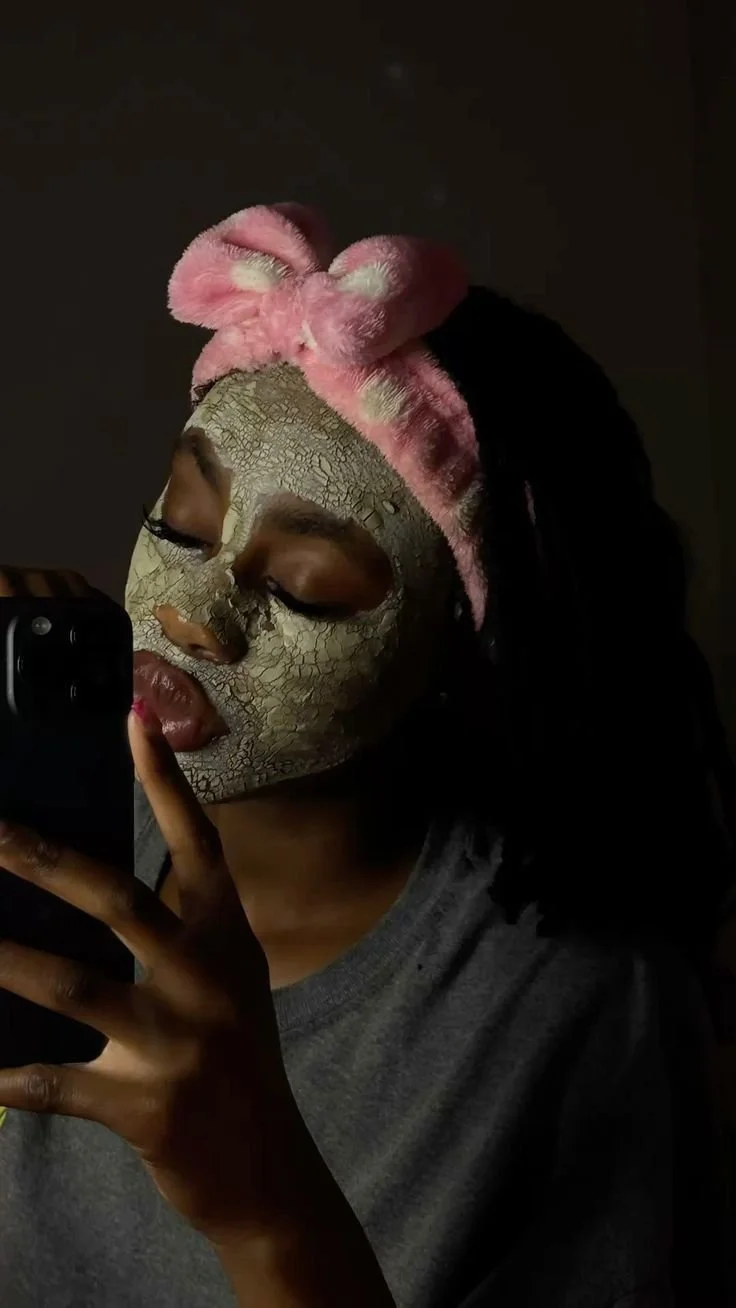 Woman applying a cracked, textured facial mask while wearing pink plush headband and taking a selfie.