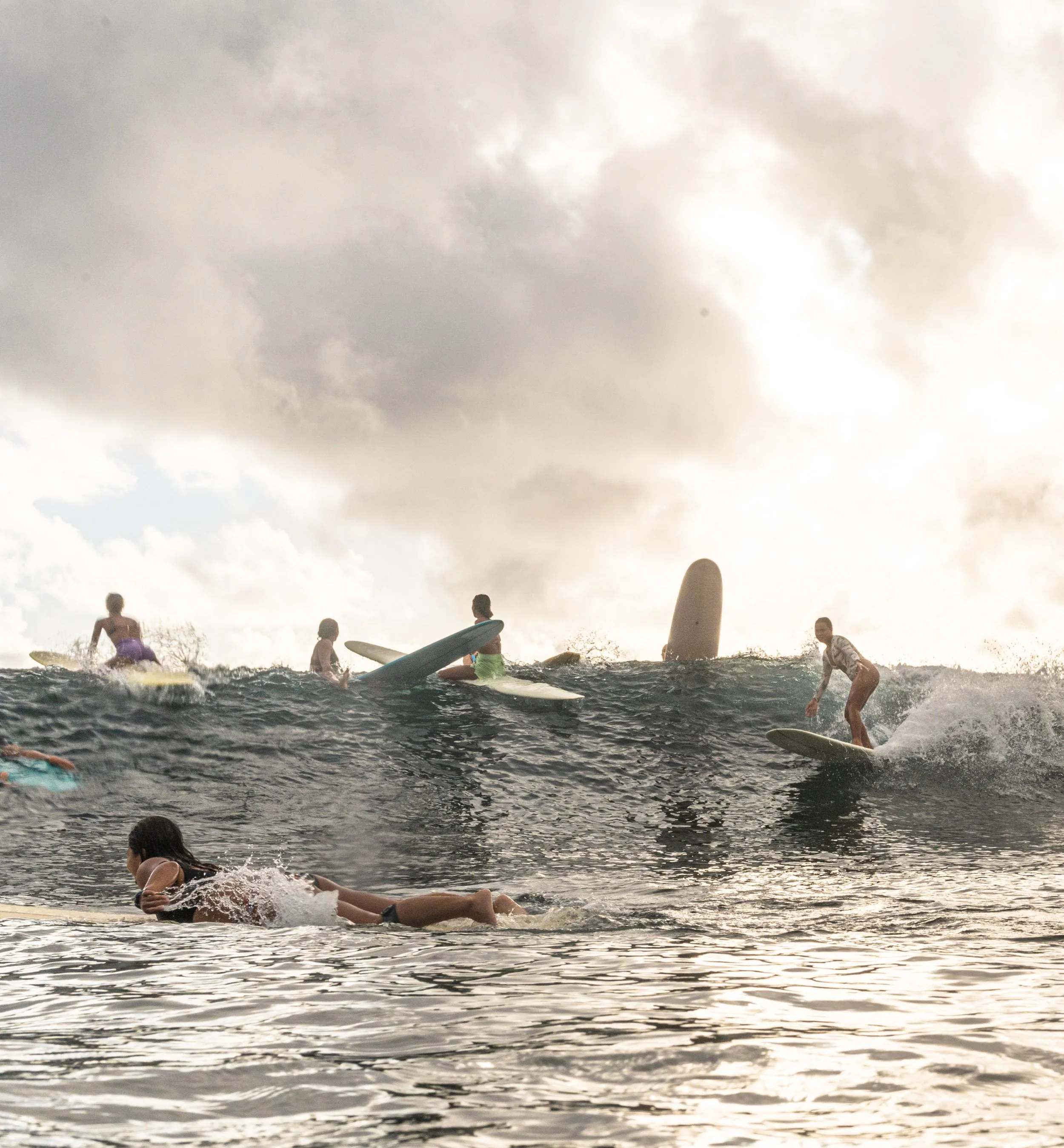 People surfing and swimming in the ocean during sunset, with cloudy sky overhead.