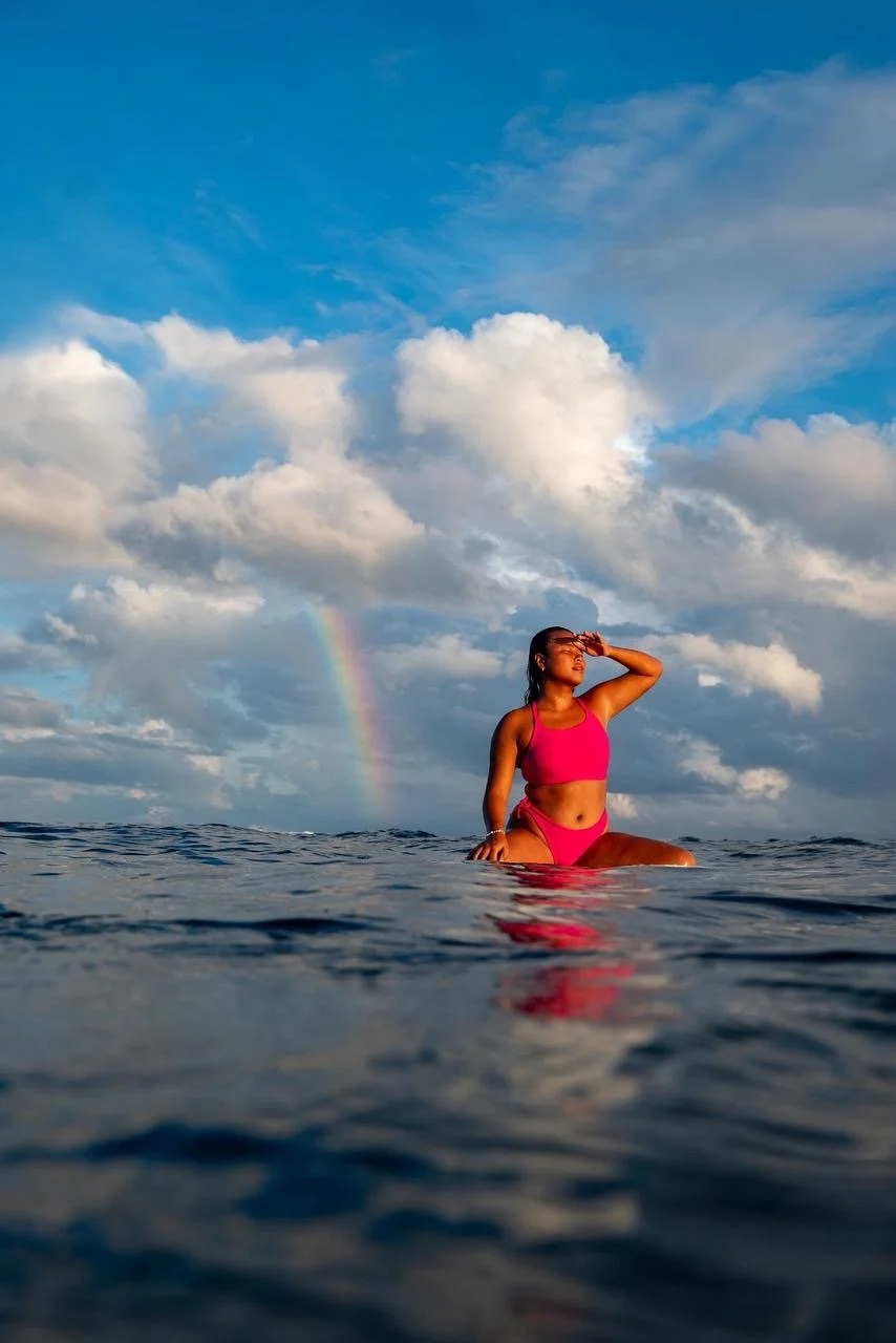 A woman in a pink swimsuit sitting in the ocean, shielding her eyes from the sun, with a rainbow and a cloudy sky in the background.