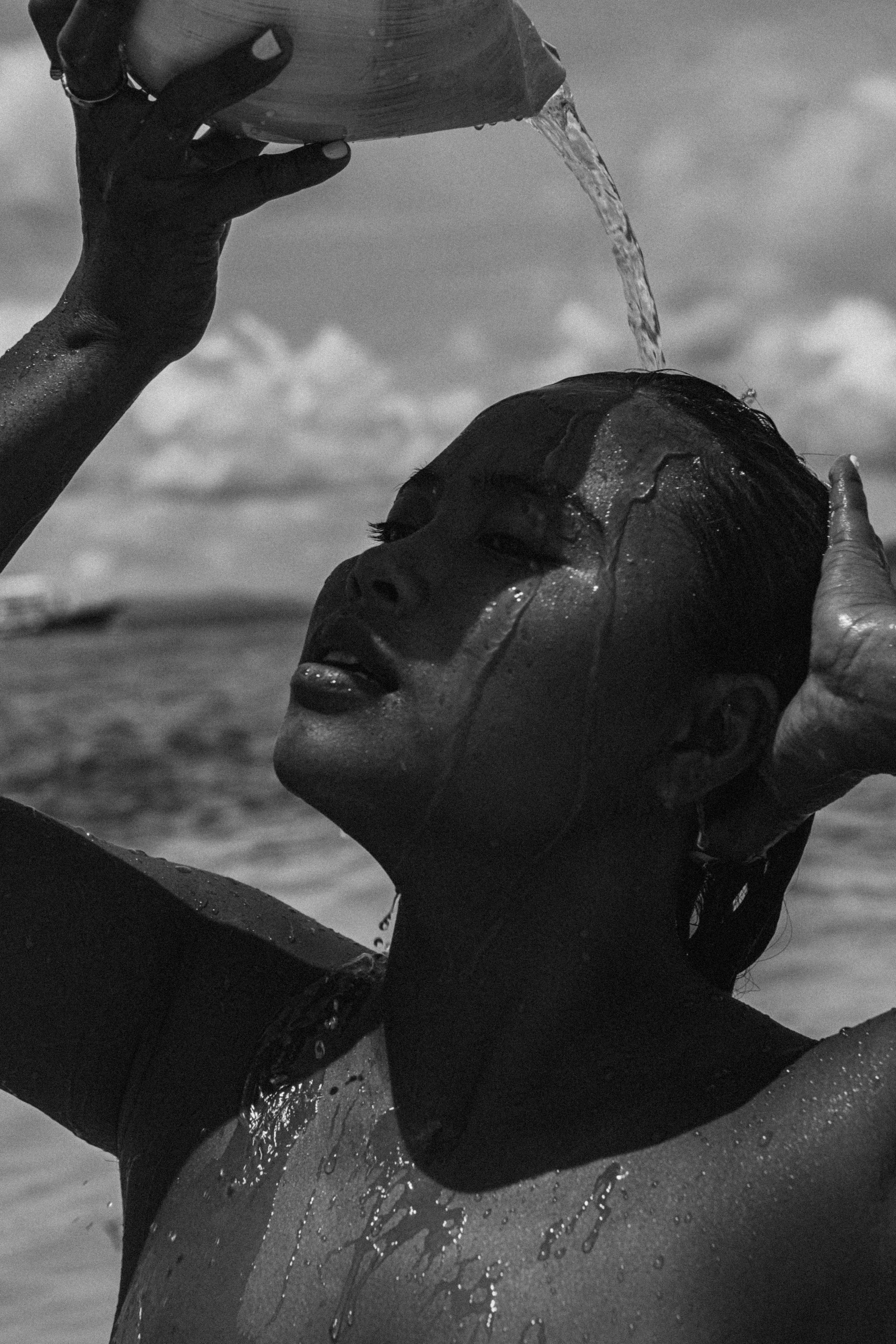 A person in a wetsuit relaxes in the ocean, with water pouring over their face and head, and a cloudy sky in the background.