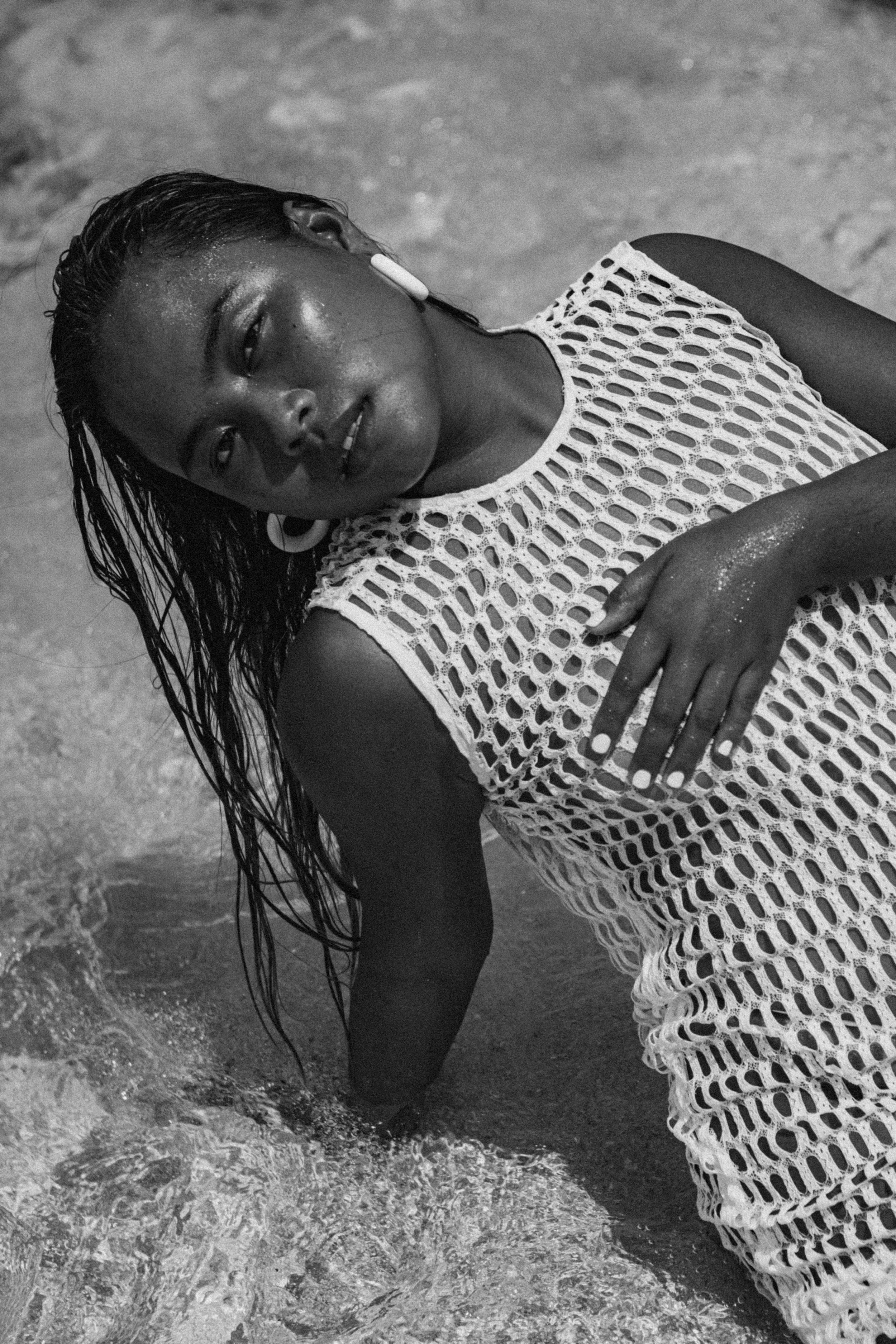 A woman lying on the beach with wet hair, wearing a mesh dress and hoop earrings, with water and sand visible around her.
