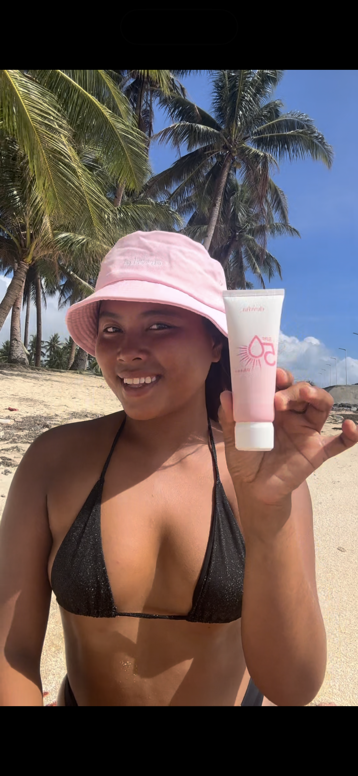 Woman at the beach wearing a pink bucket hat and black bikini, smiling while holding a pink and white tube of skincare product. Palm trees and a clear blue sky are in the background.