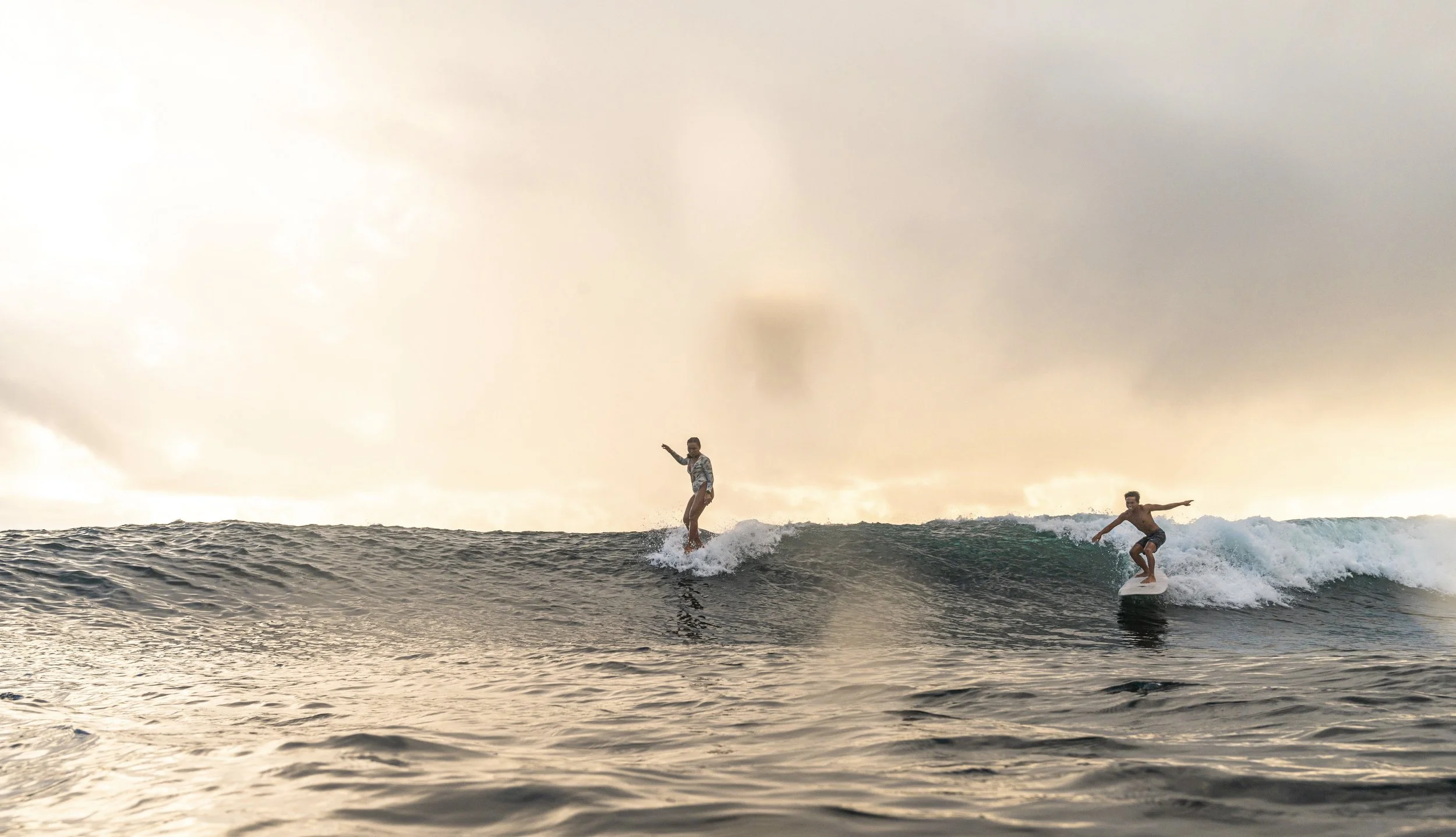 Two young men surfing on small waves during sunset or sunrise, with a cloudy sky in the background.