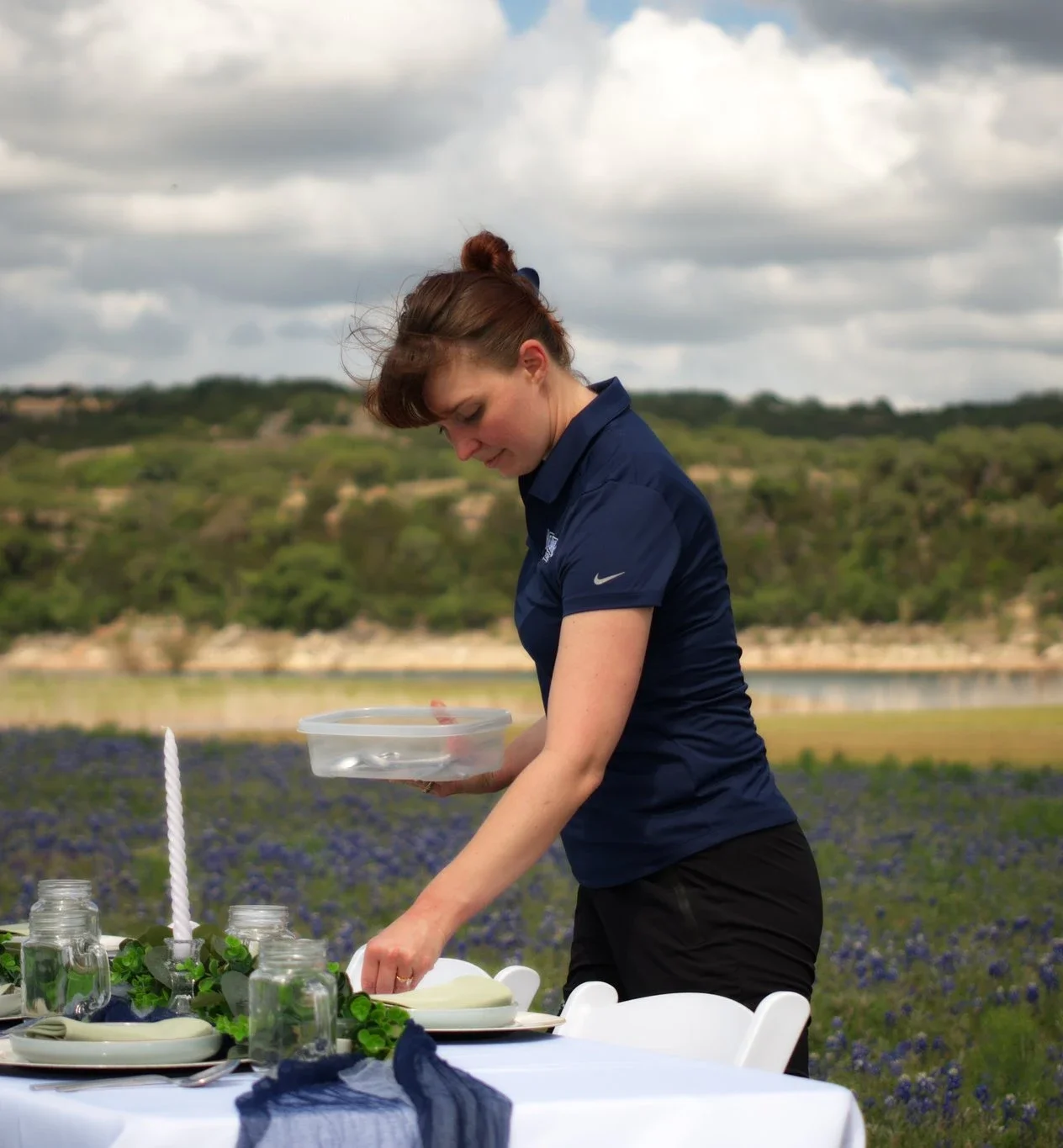 A woman setting a table outdoors in a field of purple flowers with a scenic landscape and cloudy sky in the background.