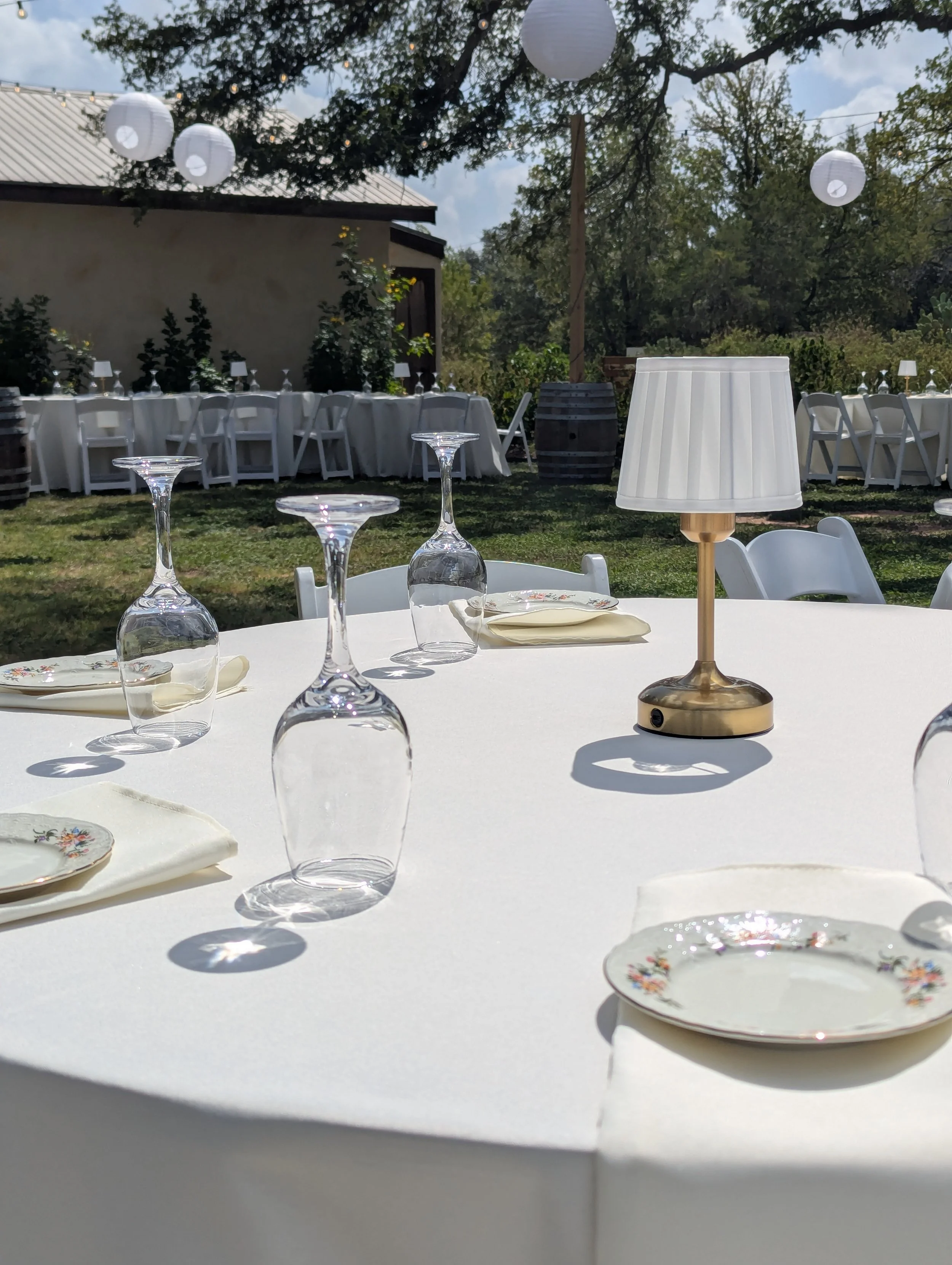 Outdoor event setup with an ivory tablecloth, glassware, floral-patterned plates, a table lamp, and hanging paper lanterns, with trees and a building in the background.