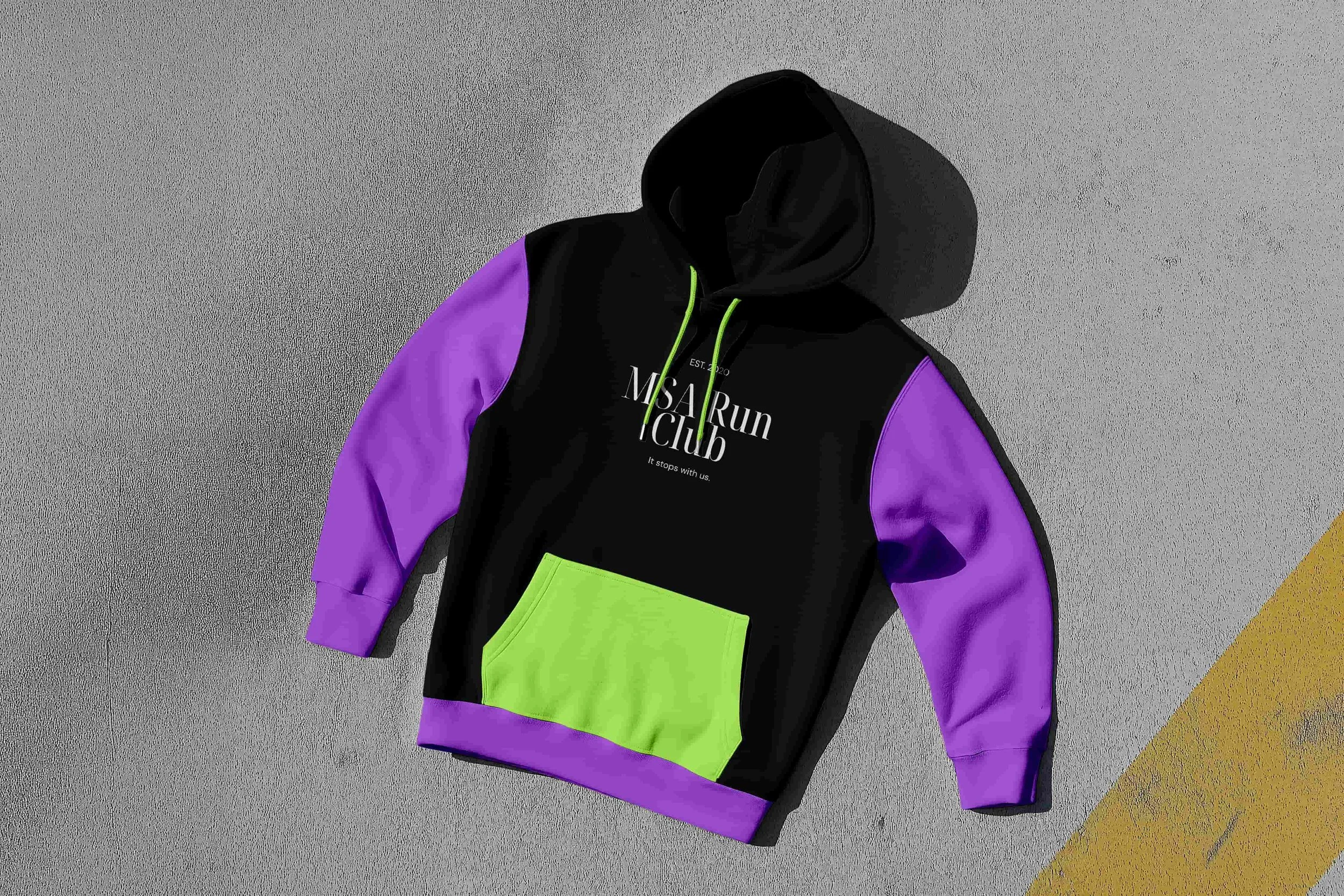 A black hoodie with purple sleeves and a neon green pocket lying on concrete ground next to a yellow line.