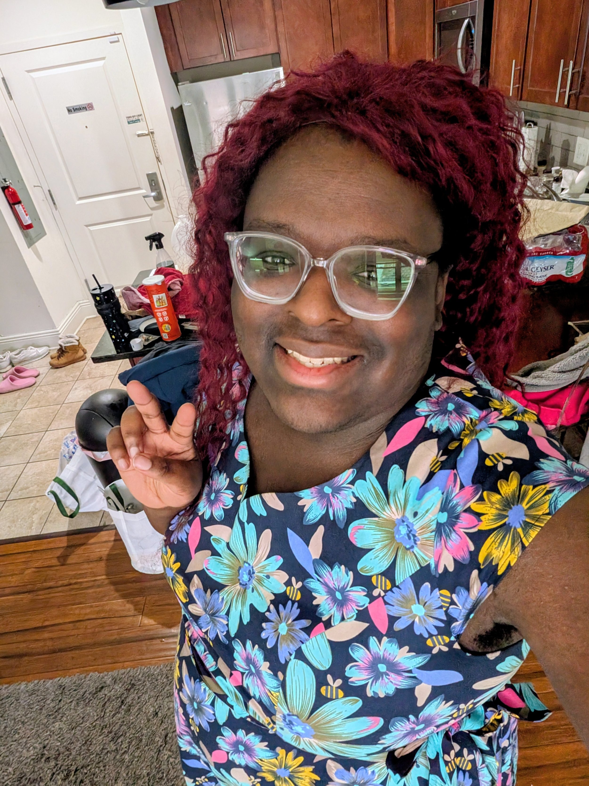 A photo of Holly, a woman with dark skin and curly red hair wearing clear glasses and a colorful floral shirt, smiling, in a kitchen with various items on the counter.