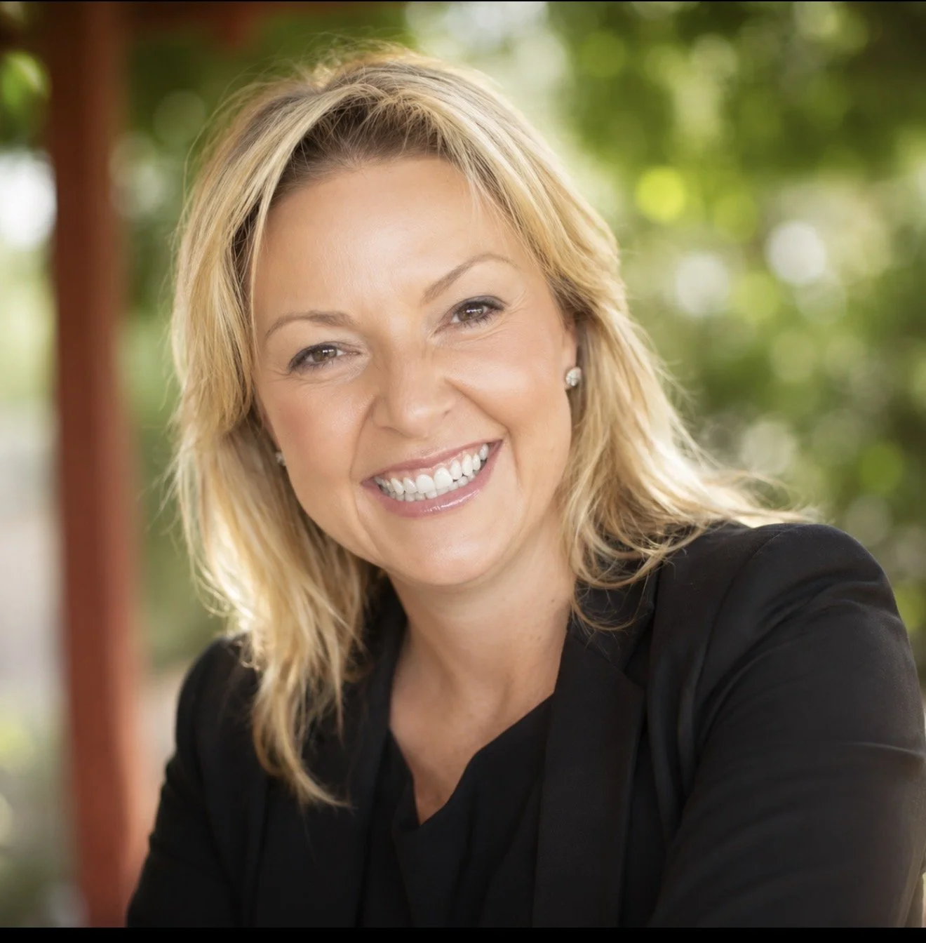 A smiling woman with blonde hair, wearing a black blazer and pearl earrings, outdoors with a green, blurred background.