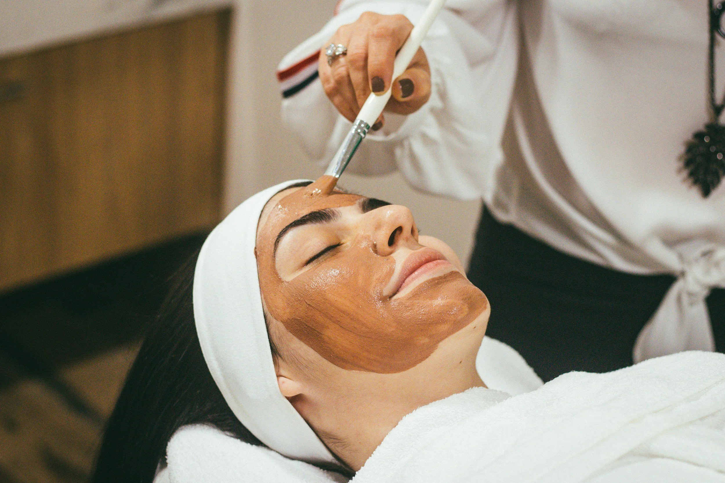 A woman is lying down with her eyes closed, receiving a facial treatment. She has a facial mask on, and a spa headband. A skincare professional is applying a product to her forehead with a brush.