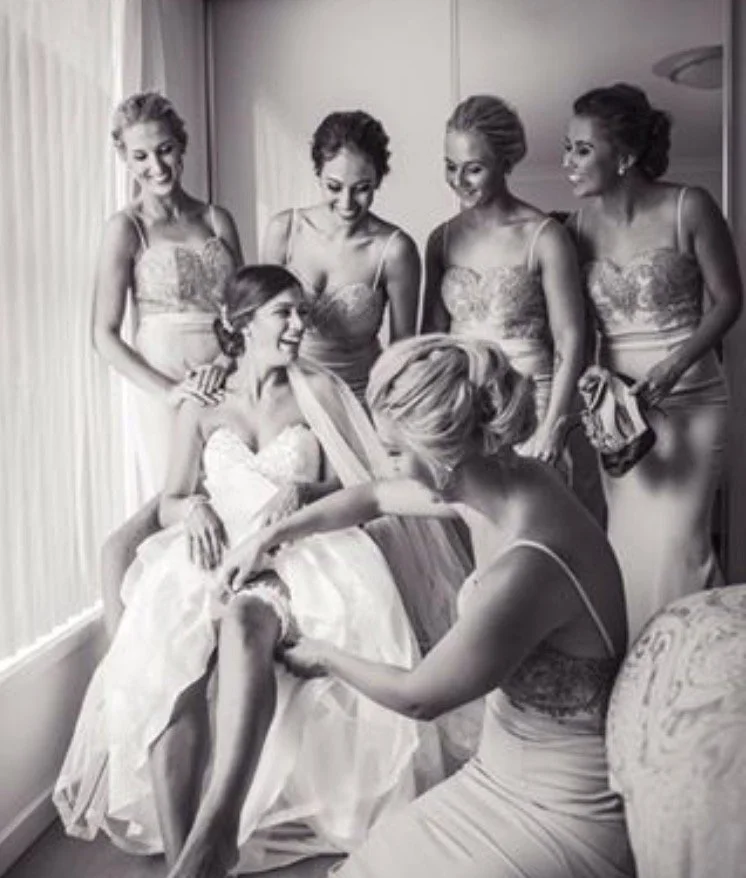 Bride in wedding dress sitting on window sill with five bridesmaids around her, one kneeling and adjusting her dress, all smiling.