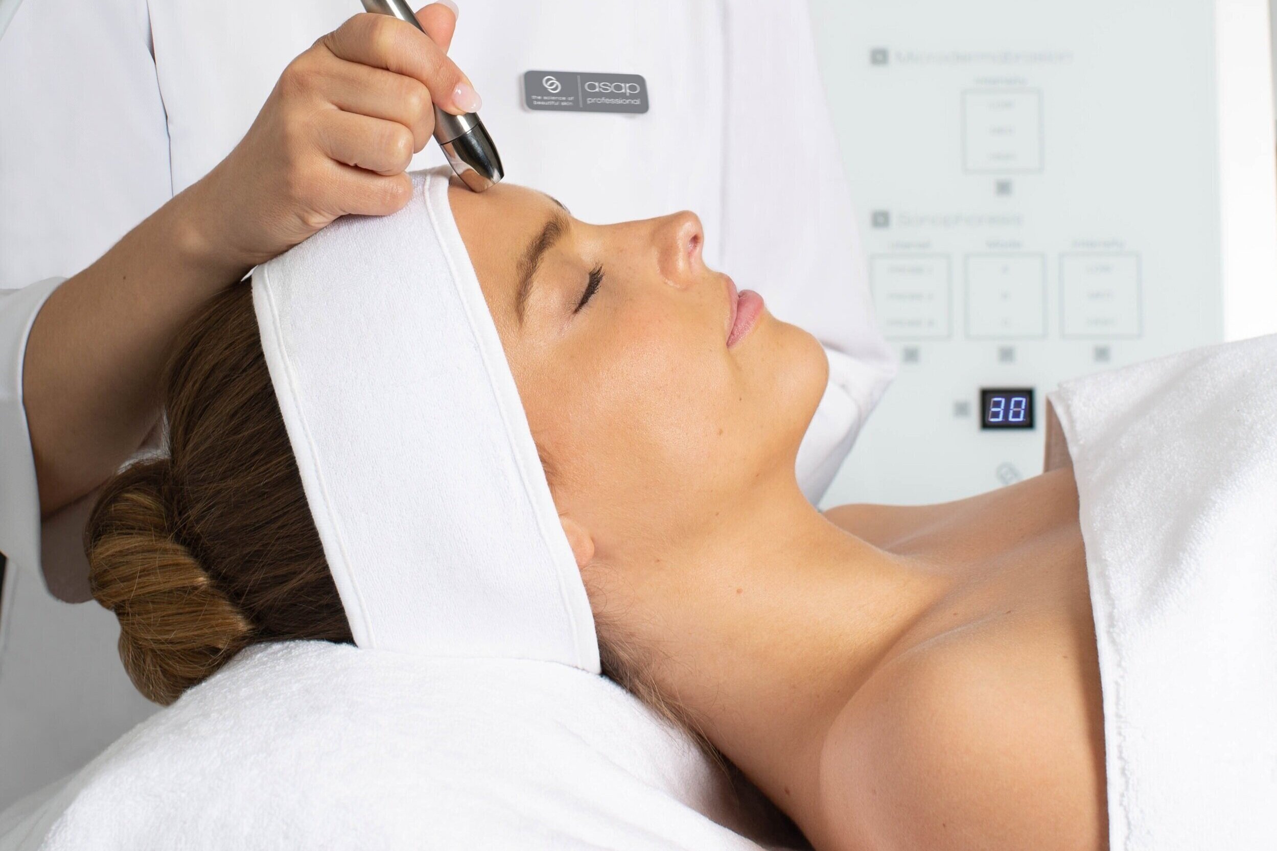 A woman lies on a treatment bed with her eyes closed, receiving a facial or skincare treatment in a spa or clinic. A professional uses a tool on her forehead.