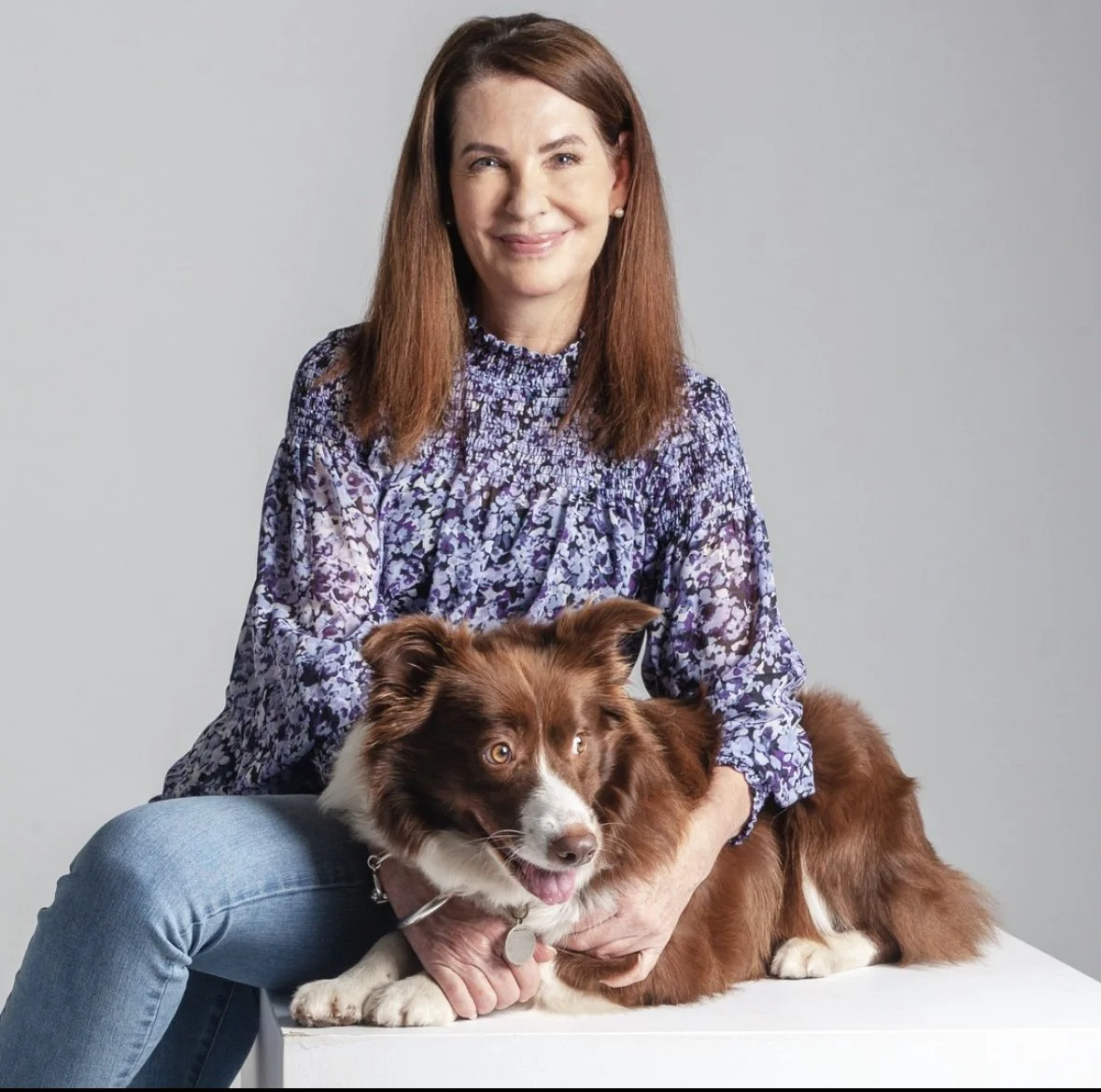 A woman with shoulder-length brown hair, wearing a purple floral blouse and jeans, sitting beside a brown and white Australian Shepherd dog with amber eyes, on a white surface against a plain gray background.
