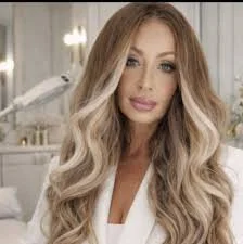 A woman with long, wavy blonde hair and makeup, wearing a white top, in a well-lit bedroom or dressing room.