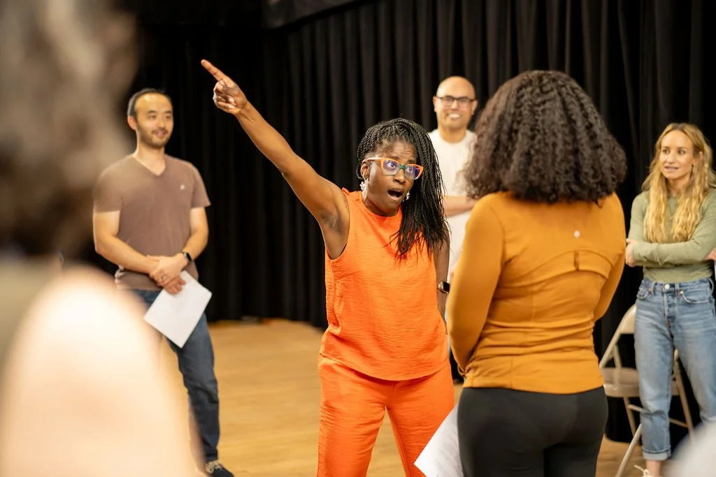 A student in orange points emphatically up in the air, making a dramatic point to her scene partner while classmates look on, in a Bethesda Improv Theater class.