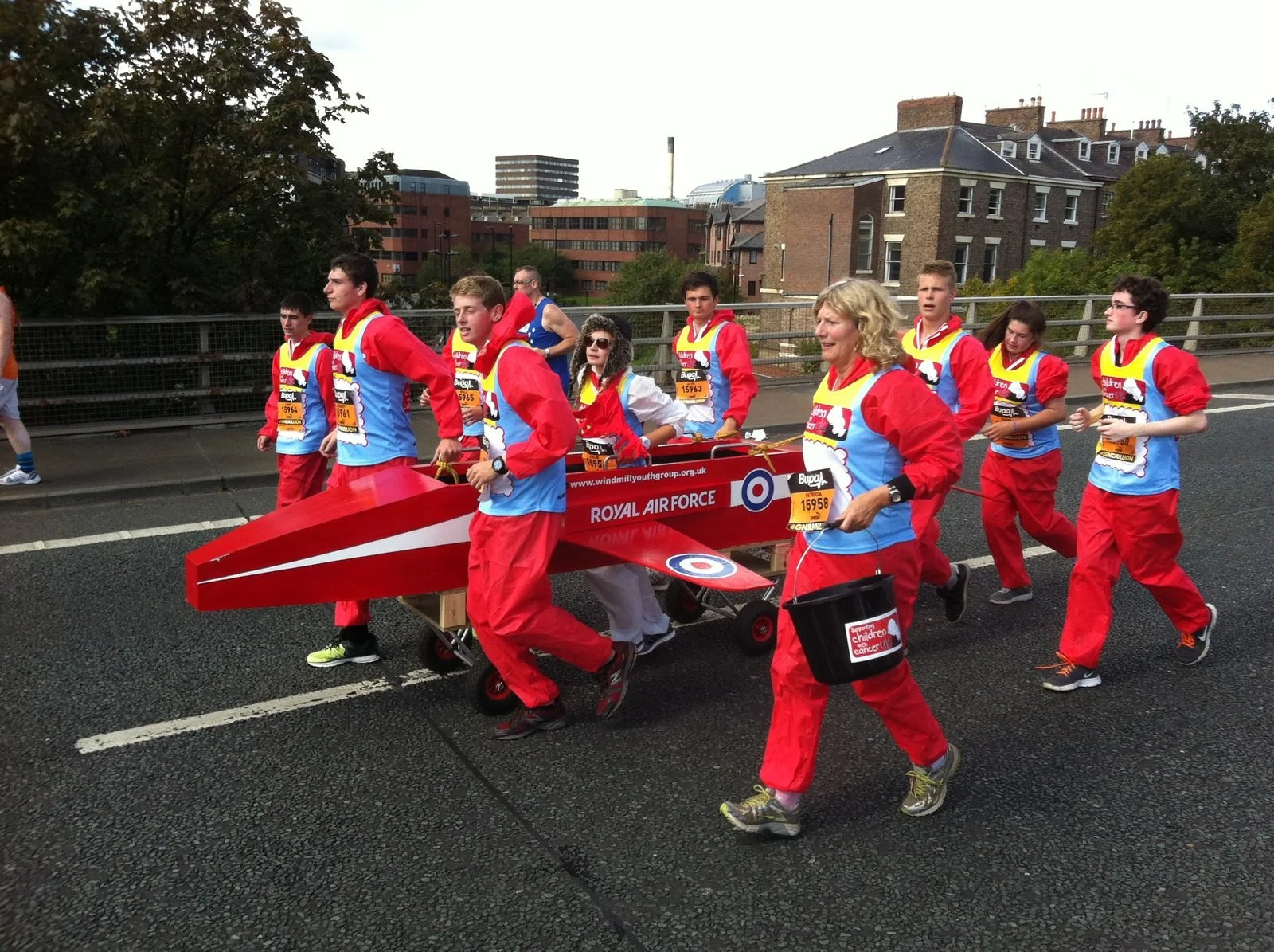 Group of people dressed as Royal Air Force airmen pushing a model airplane on a street during a parade or event.