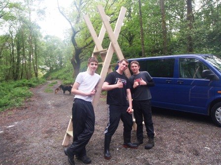 Three young men standing outdoors near a blue van and a large wooden structure, with trees and a dirt path in the background.