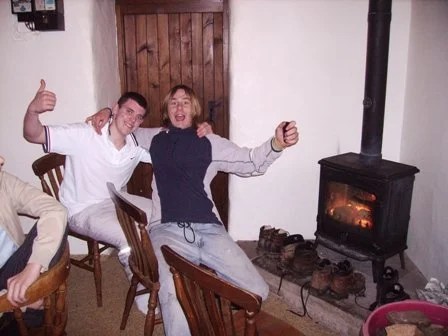Two young men sitting on chairs near a wood stove, smiling and gesturing with their hands, in a cozy room with a wooden door and a white wall.