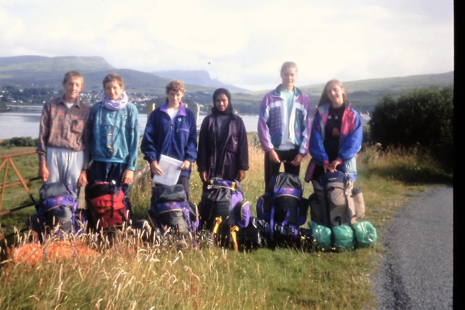 Group of six children with backpacks standing outdoors by a lakeside with hills in the background.