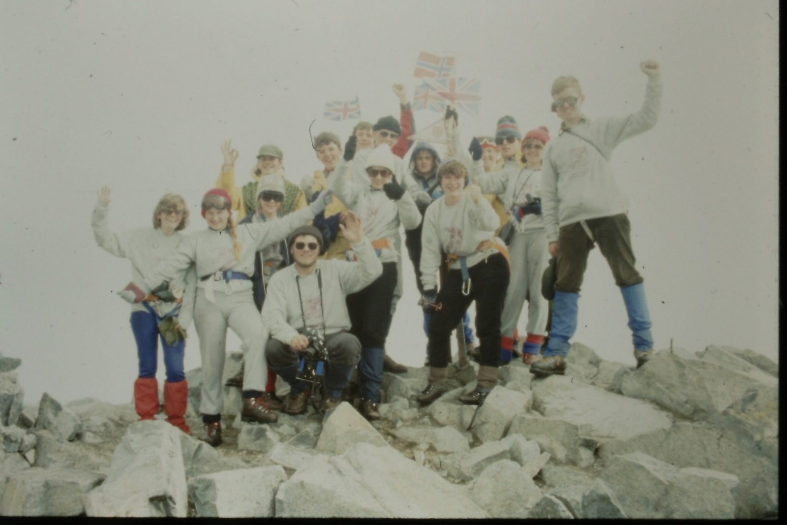 Group of people on a rocky summit celebrating with British flags, dressed in outdoor gear and winter clothing.