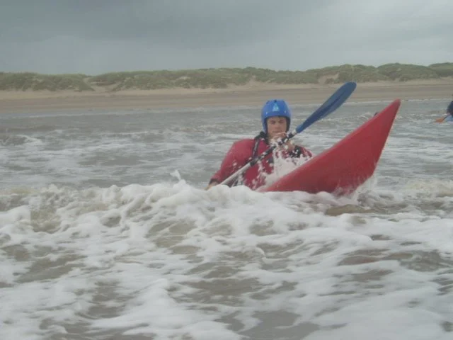 Person in a red kayak paddling through ocean waves, wearing a blue helmet and life jacket.