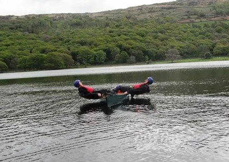Two people wearing life jackets and helmets in a small boat on a lake, with green hills in the background.