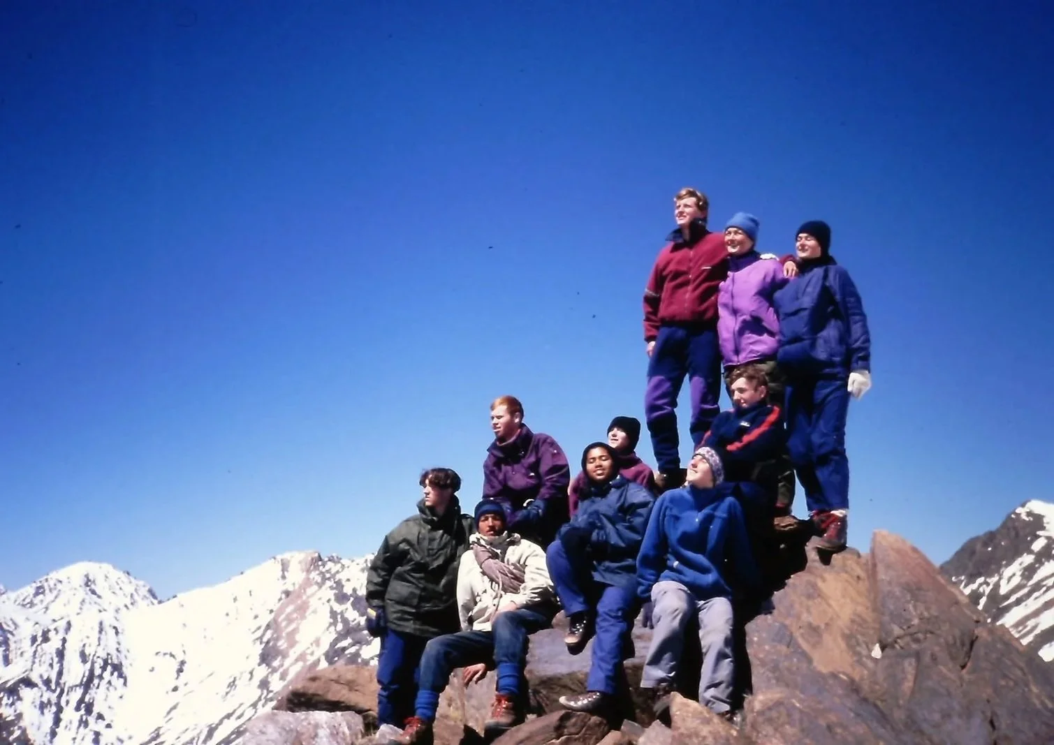 Group of hikers in winter clothing posing on a mountain peak with snow-capped mountains in the background.