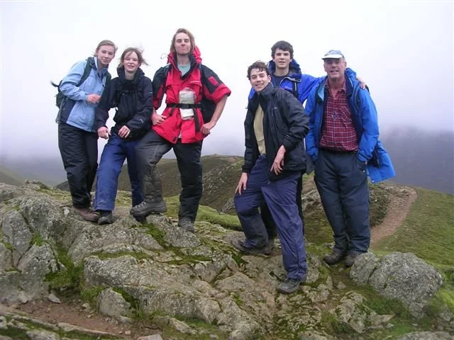 Six hikers in outdoor gear standing on a rocky trail with grassy hills behind them, foggy weather, and a cloudy sky.