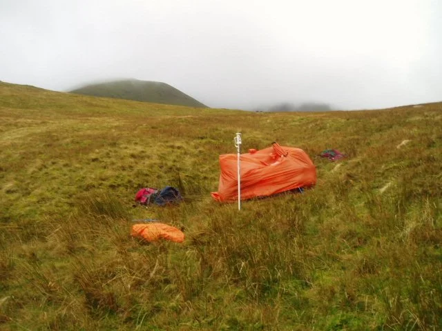 A camping tent and gear set up on a grassy hillside with mountains in the background, under overcast sky.