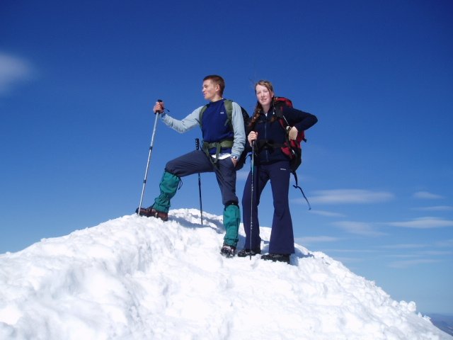 Two hikers standing on a snow-covered mountain peak under a clear blue sky, holding hiking poles and wearing backpacks.