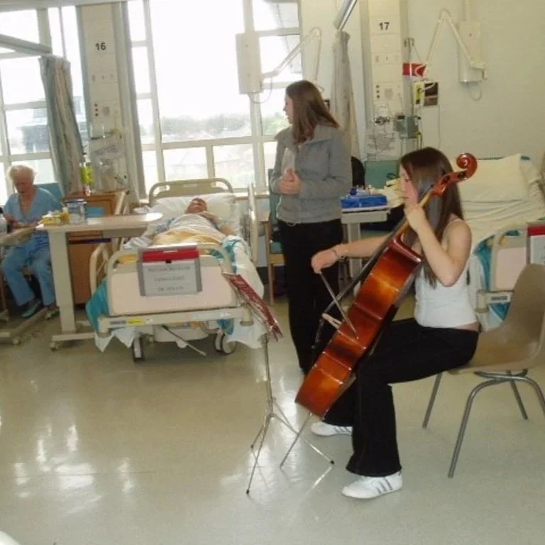 A young woman playing the cello in a hospital room while a nurse and a patient are visible in the background.