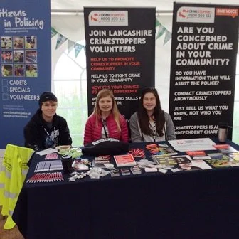 Two women sitting at a table promoting Lancaster Crime Stoppers, with banners about volunteering and community crime concern behind them.