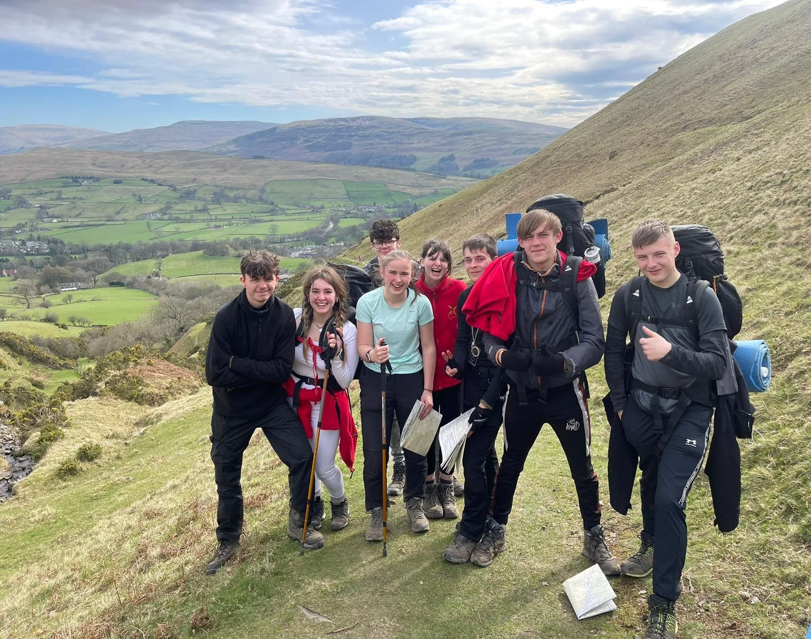 Group of young hikers smiling and posing on a grassy hillside with a scenic countryside in the background, carrying backpacks and walking gear.