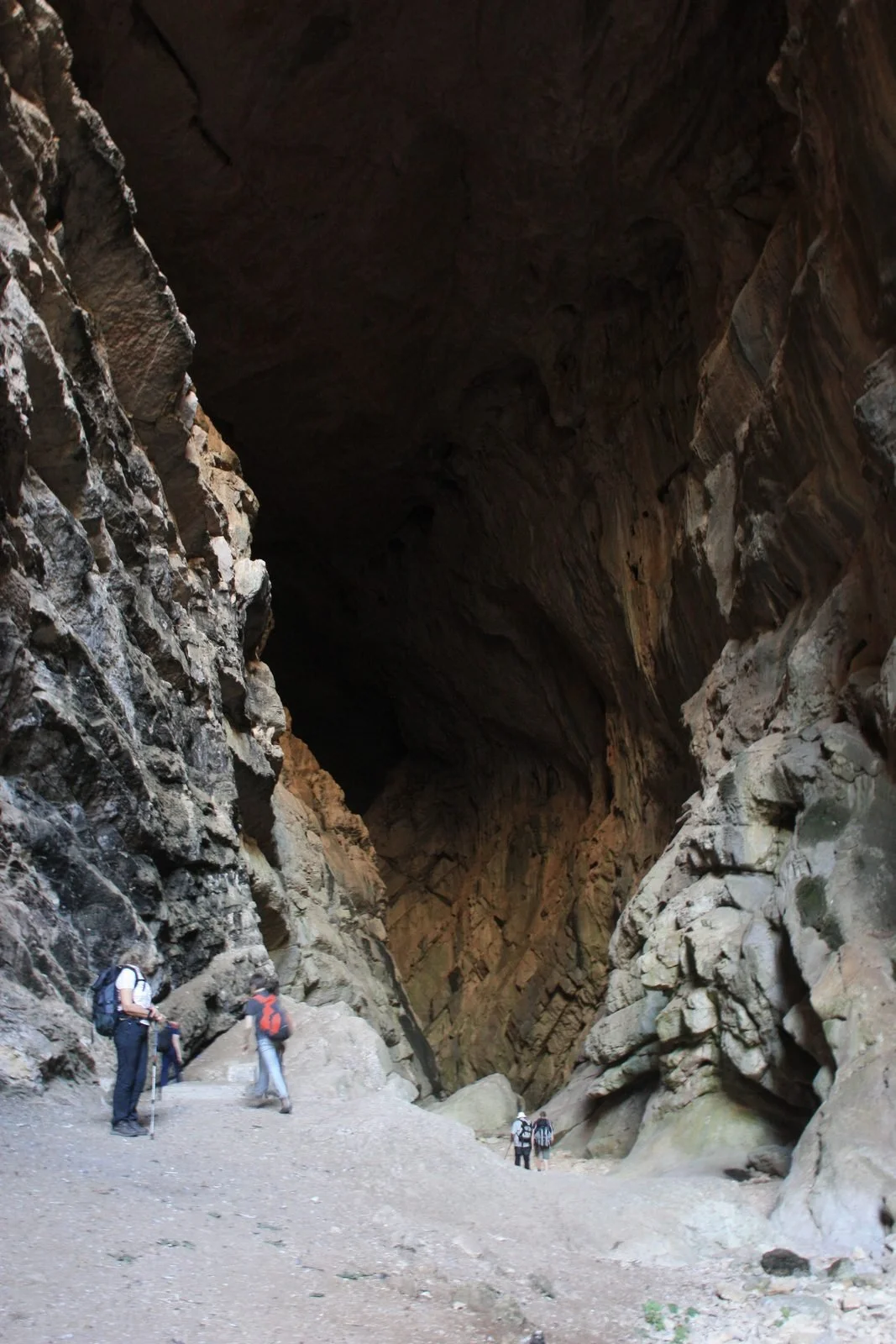 Hikers walking in a large, rocky cave with high, dark ceiling and rugged walls.