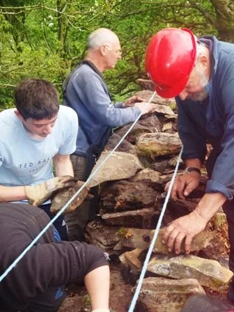 Three people working together to build or restore a stone wall outdoors, one wearing a hard hat, in a wooded area.