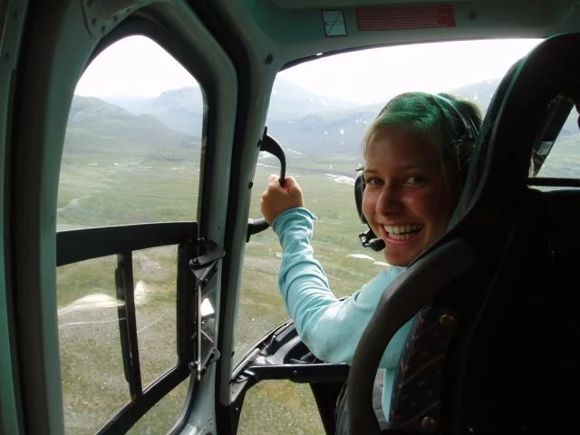 A woman smiling in the cockpit of a helicopter, holding the control stick, with mountains and a valley visible outside the window.
