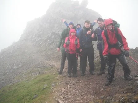 Group of five hikers on a mountain trail, dressed in outdoor gear, with rocky mountain and fog in the background.