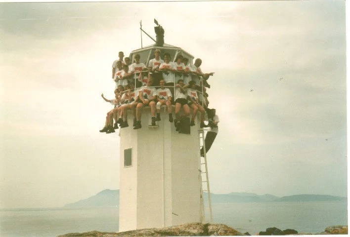Group of children sitting around the top of a lighthouse, with one standing and waving a flag, overlooking the ocean and distant islands.