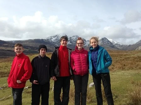 Five people standing outdoors in front of mountains with snow, cloudy sky, and grassy terrain.