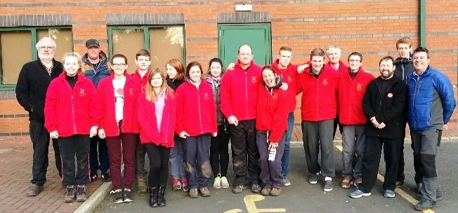 Group of people standing outdoors in front of a brick building, many wearing red jackets, some with their arms around each other.