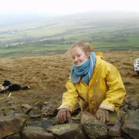 A woman in a yellow rain jacket and blue scarf leaning on rocks outdoors, smiling, with a grassy landscape and distant hills in the background.