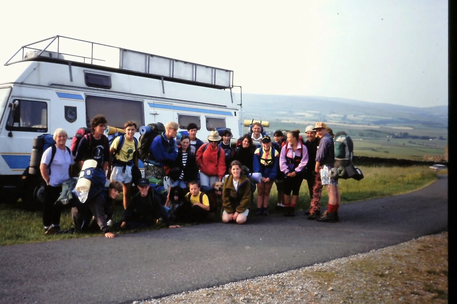 Group of people with backpacks and gear standing in front of a white van in a rural area with rolling hills in the background.