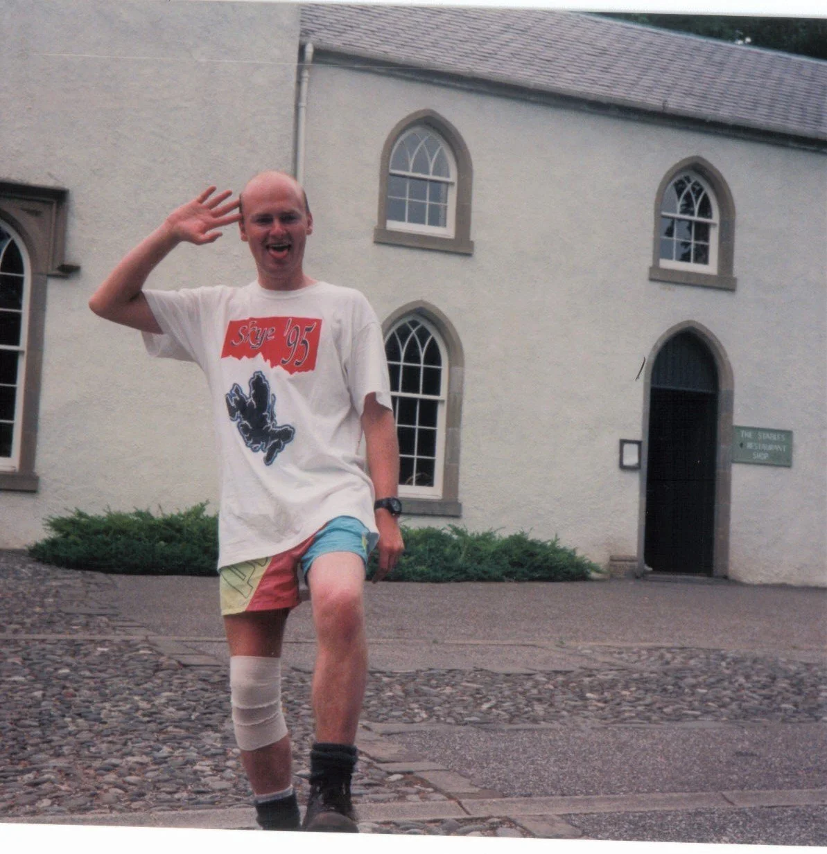 A man smiling and waving while standing on a cobblestone street in front of a white building with arched windows. He is wearing a white T-shirt, colorful shorts, black boots, and has a beige cast on his left knee.