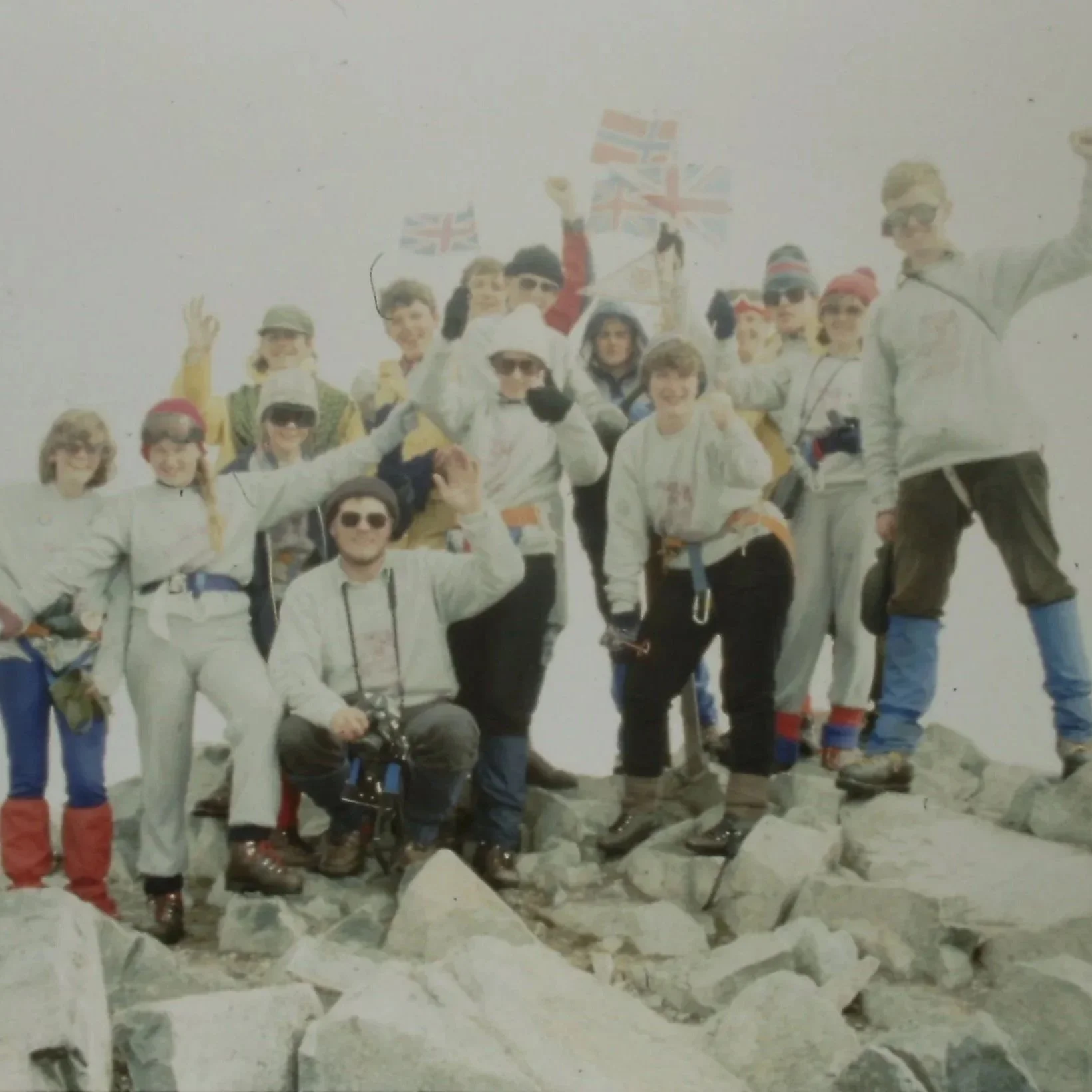 Group of hikers at mountain summit with flags, rugged terrain, and rocky ground.