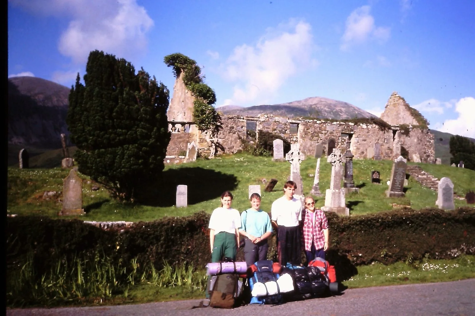 Group of four people standing in front of an old stone church and graveyard, with mountains in the background and luggage on the ground.