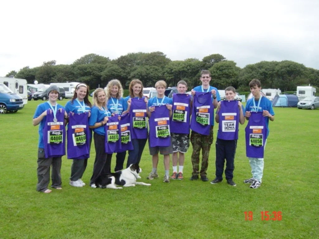 Group of eight children with medals and purple bags, standing on green grass, with a dog in front, at an outdoor event with tents and vehicles in the background.