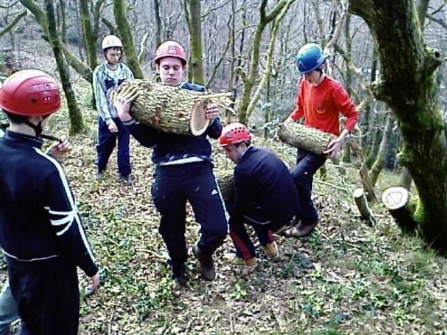 Group of young people wearing helmets and outdoor clothing in a wooded area, carrying and balancing logs.
