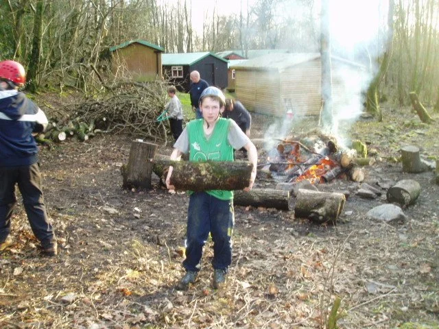 Children and adults outdoors gathering around a campfire, with one boy carrying a large log in foreground, trees, sheds, and smoke rising from the fire in the background.